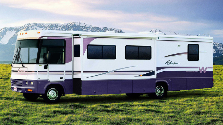 A large, modern Winnebago with purple accents and the slide pushed out sitting on grass with snowy mountains in the background