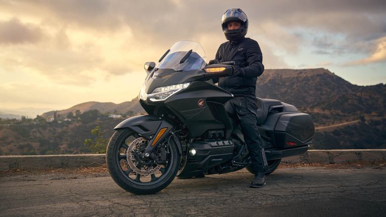 A rider in full kit and helmet sitting atop a stationary Honda Gold Wing motorbike finished in black metallic.