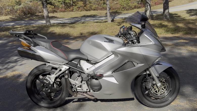 A beautiful chrome silver Honda VRF800 Sport tourer bike parked outdoors in the shade of some trees on a road.