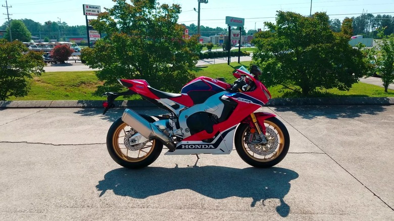 A 2017 Honda Fireblade SP CBR1000RRR standing on pavement, with a main road and some plants in the backdrop.