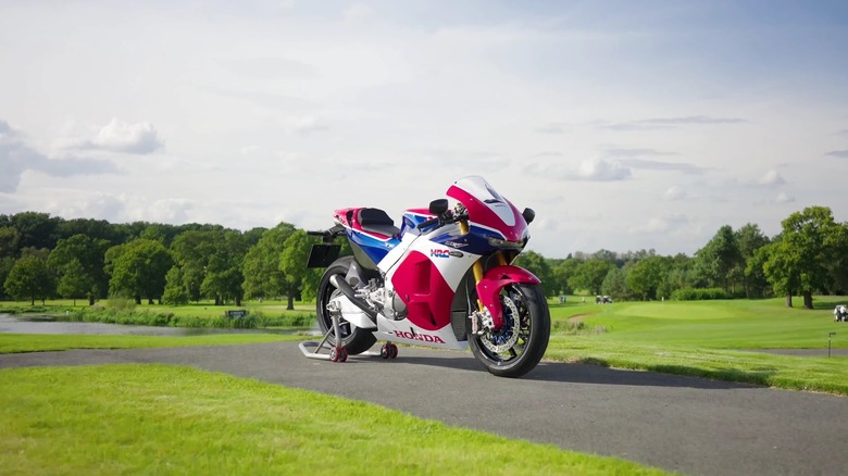 A red, white, and blue Honda RC213V-S street legal hyperbike standing on a roller rear wheel stand on a smal road, surrounded by grass.