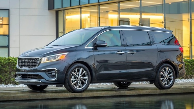 A metallic grey Kia Sedona 2021 against the backdrop of a glass building, parked horizontally near a water feature
