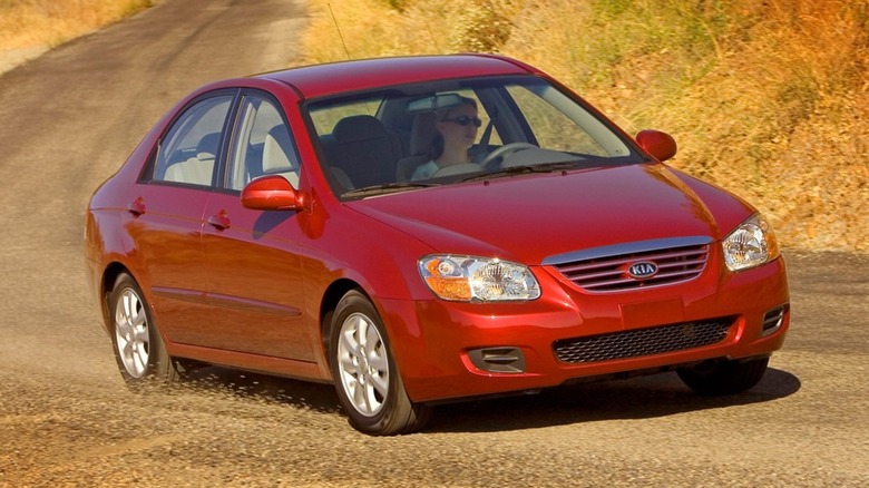 A blue 2007 Kia Spectra against a forested background, parked diagonally on a road.