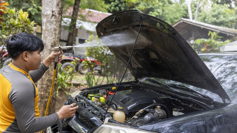 A focused man doing car maintenance outdoors by pressure washing the engine bay of a vehicle.