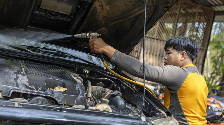 A car mechanic in overalls going about pressure washing the engine bay of an unknown car.