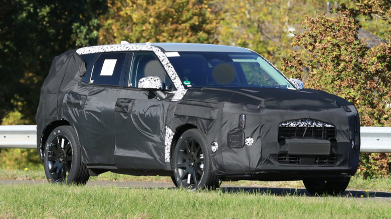 202X Kia Sportage test vehicle in camouflage and partly covered with tarps driving on road, front-right 3/4 view