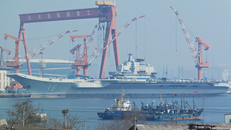 The Liaoning, a Chinese aircraft carrier in dock with massive naval shipyard cranes and derricks in the background