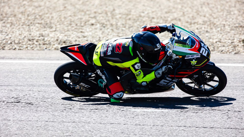 Racer in full Aprilia Racing gear cornering aggressively on an Aprilia RS 250 SP track bike, with the rider's knee nearly touching the asphalt during a high-speed lean.