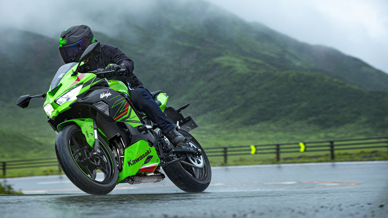 Rider leaning into a turn on a bright green Kawasaki Ninja ZX-25R sportbike in wet conditions, with misty green mountains and a wet road surface in the background.