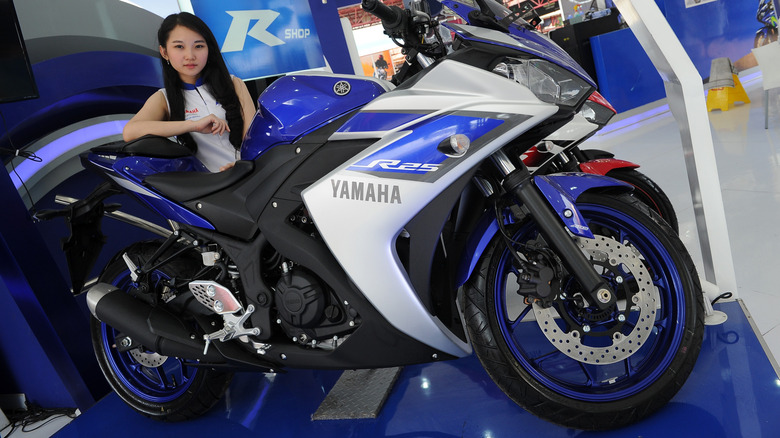 Yamaha R25 sportbike in blue and silver on display at an indoor showroom, with a promotional model posing beside the motorcycle and Yamaha branding visible in the background.