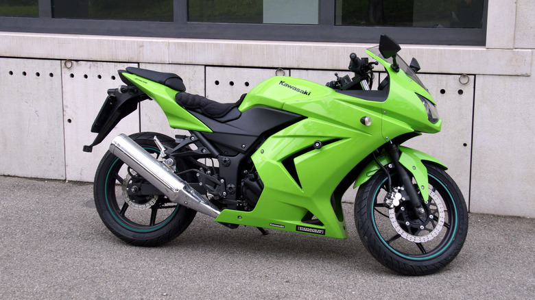 Side view of a Kawasaki Ninja 250R sportbike in lime green, parked on pavement in front of a modern concrete building with dark windows.