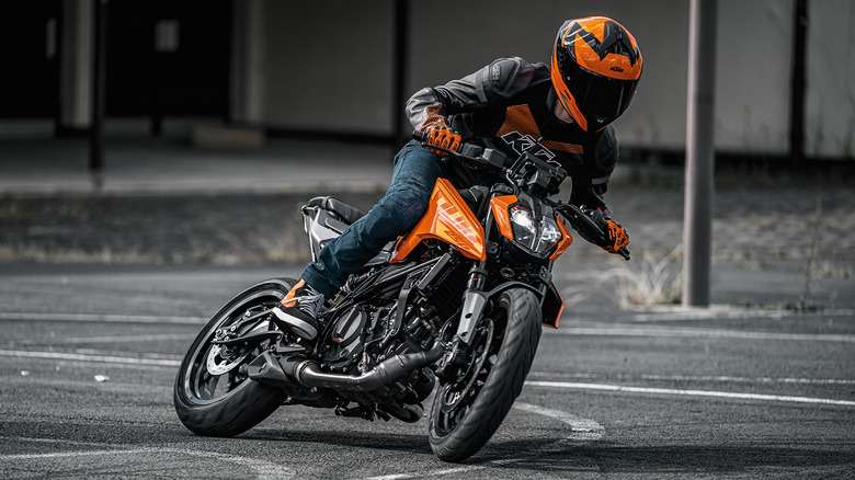 Rider in full KTM-branded gear leaning into a sharp turn on a bright orange KTM Duke 250 motorcycle, captured mid-action on an empty asphalt lot.