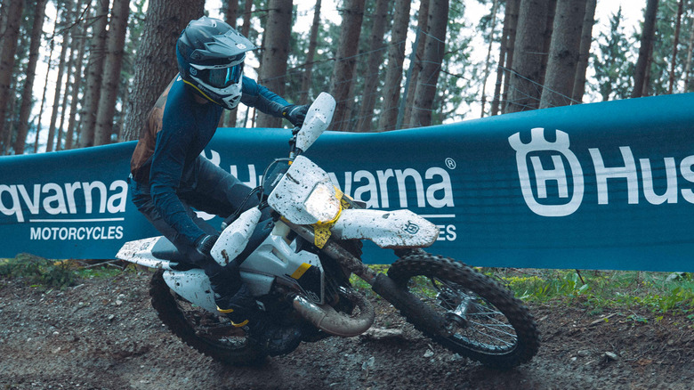 Rider navigating a dirt trail on a Husqvarna TE 250 enduro motorcycle during an off-road race in a forest setting, with Husqvarna Motorcycles branding in the background.