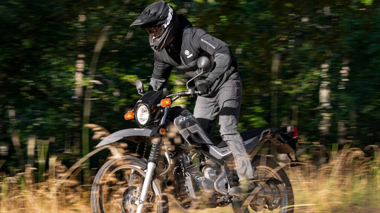 Motorcyclist wearing full gear and a black helmet riding a Yamaha XT250 off-road through tall grass, with motion blur in the background indicating speed and movement.