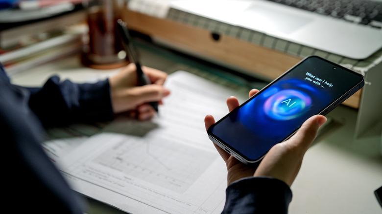 A person sitting a desk writing with one hand while holding a smartphone with an open AI app in the other hand