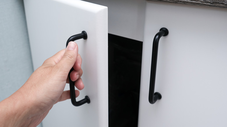 A hand opening a white kitchen or bathroom cabinet with black handles