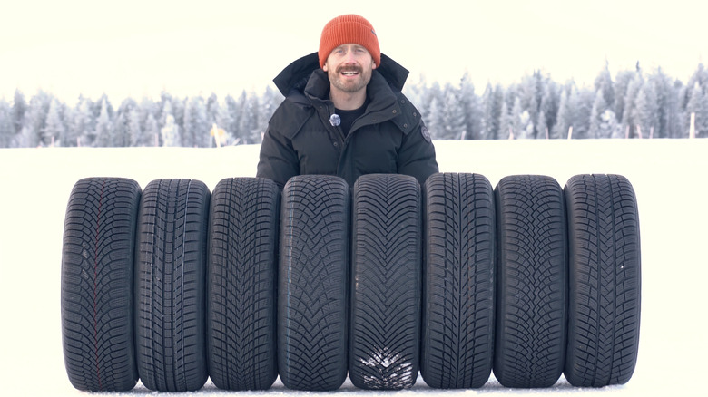 A TyreReviews tire reviewer standing behind a row of winter tires in snowy conditions.