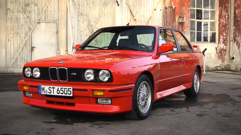 A bright red BMW M3 (E30) with BBS-style mesh wheels and an M badge on the grille is parked in front of a weathered industrial building with red paint peel-offs.