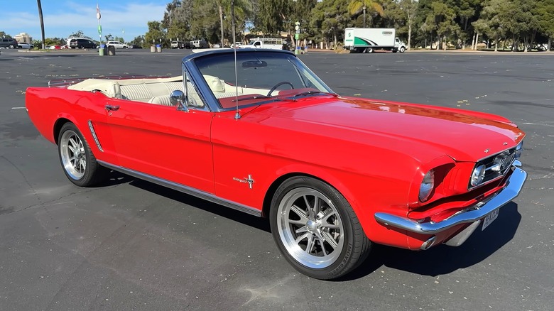 A 1965 bright red Ford Mustang convertible with its top down and cream interior parked in a sunny open lot lined with trees.