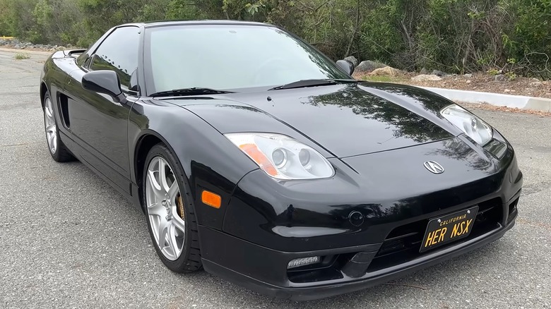 A 2005 black Acura NSX with a custom California vanity plate reading "HER NSX" parked on a road surrounded by dense brush.