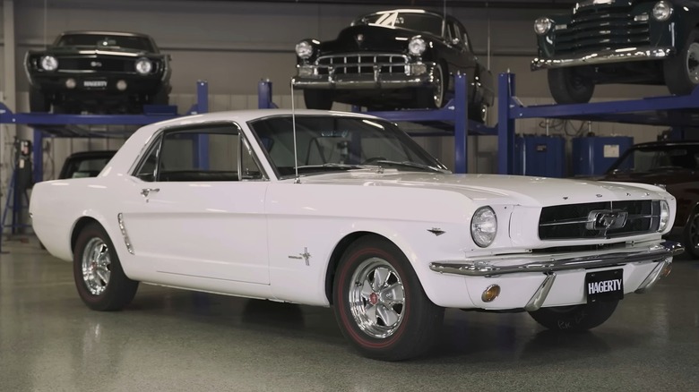 A white first-gen Ford Mustang coupe with chrome five-spoke wheels, red-stripe tires, and a Hagerty license plate frame sitting on a polished garage floor surrounded by several classic cars stacked on blue four-post lifts in the background.