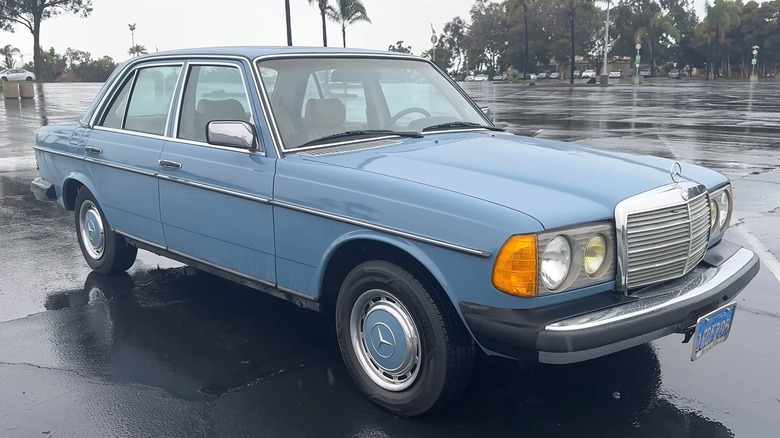 A powder blue Mercedes-Benz W123  300D sedan with chrome trim, steel hubcaps, and amber fog lights sitting in a wet parking lot on an overcast day.