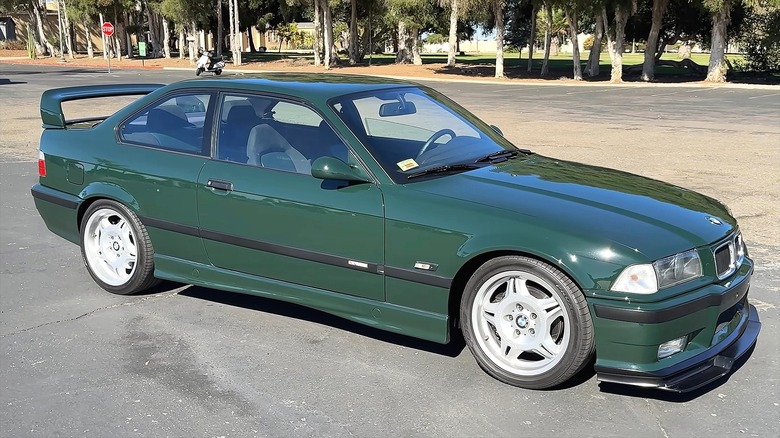 A British Racing Green BMW E36 M3 with white M-style five-spoke wheels and a rear spoiler parked in a sunny lot surrounded by tall trees.