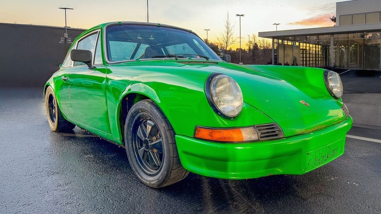 A photo of a 1973 vibrant lime green early-gen Porsche 911 with wide fenders, round headlights, and black Fuchs-style wheels being photographed at dusk in a wet parking area.