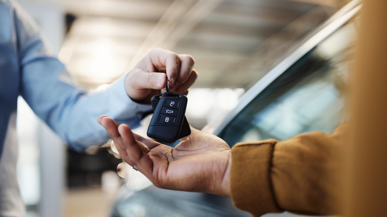 A person handing another person a set of car keys, out of focus car in the background