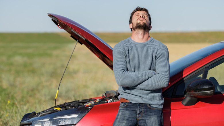 A frustrated man in a grey sweater and jeans leaning against a red car broken down on the road with its hood raised