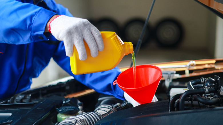 A mechanic in blue coveralls and white gloves pouring oil into a car engine with a red funnel