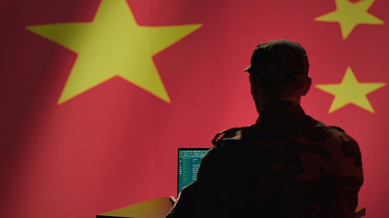 A person in uniform sits at a laptop, with the Chinese flag prominently visible in the background.