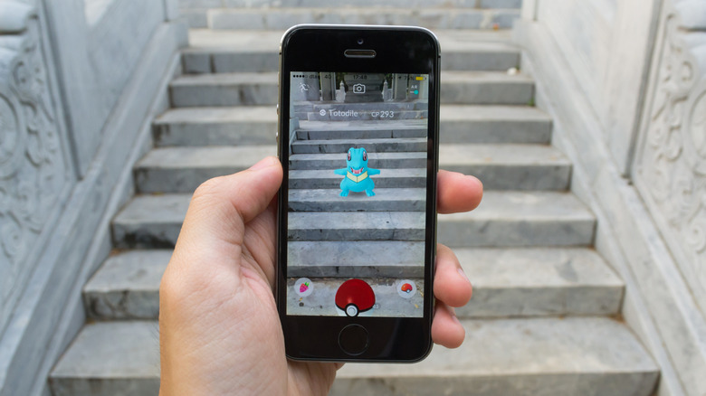 A hand holding a smartphone playing Pokemon Go in front of some gray stone steps. On the screen is an image of the same steps with a Pokemon in the center.