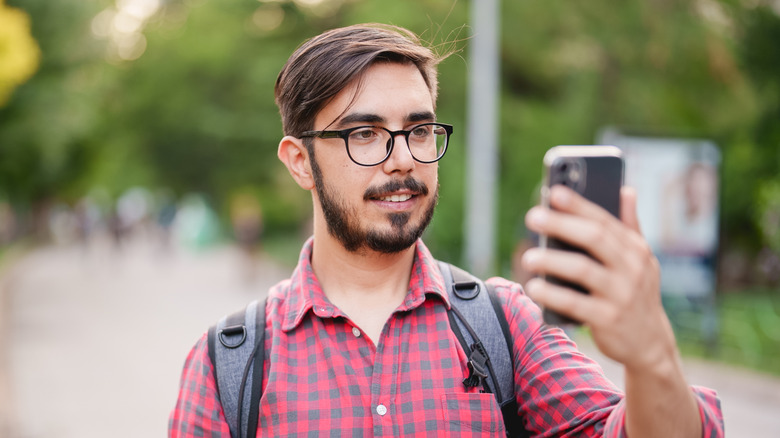 A man with glasses, a plaid shirt and a backpack is holding up a smart phone as though recording a video. The background is blurred but shows an outdoor scene.
