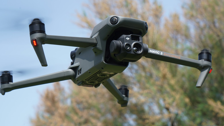 A camera-equipped drone hovering with trees in the background.