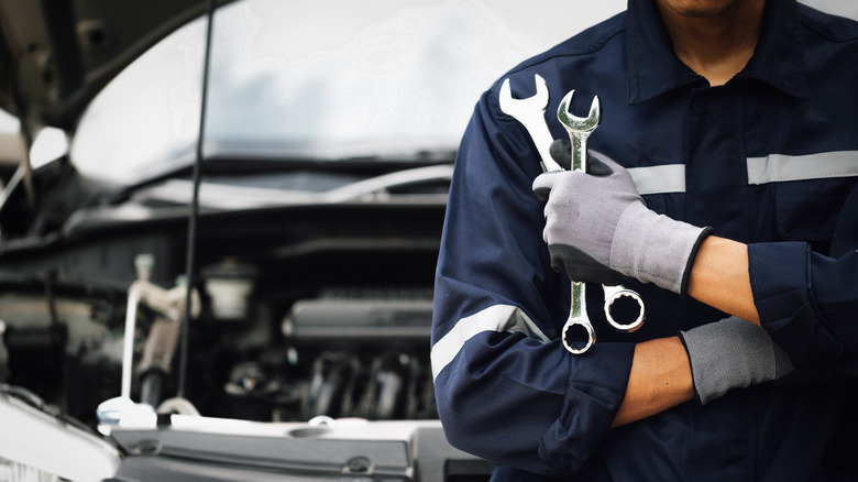 An auto mechanic holding two wrenches in front of open car hood