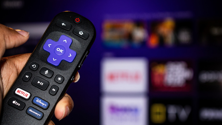Close-up of a hand holding a Roku TV remote with a blurred Roku menu displayed in the background
