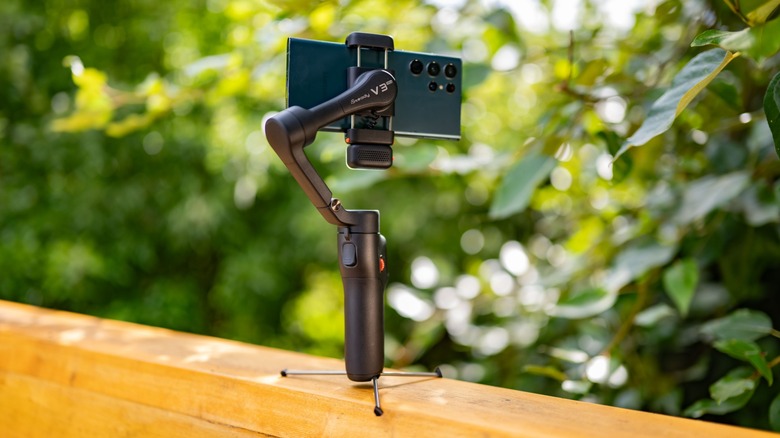 The Hohem iSteady V3 Ultra with its tripod deployed on a fence railing carrying a smartphone.