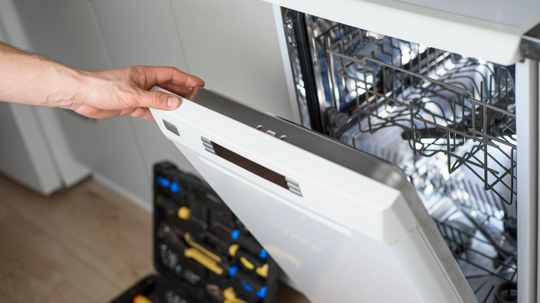 A person's hand is opening a dishwasher's door with repair tools seen next to it.