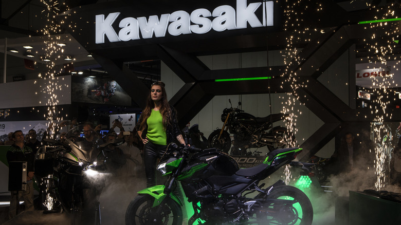 A Kawasaki Versys 1000 motorbike being revealed at a show in the dark under a lit Kawasaki logo.