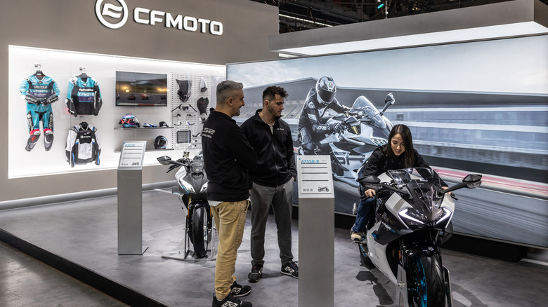 A CFmoto stand and podium finished in gray at an indoor trade event, with people looking at their offerings.