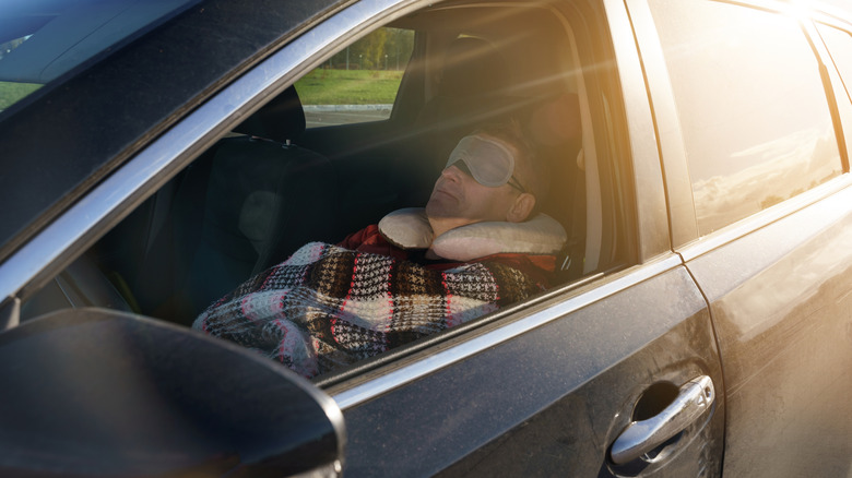 A tired driver sleeps in the car during the day, covered with a blanket and wearing a sleep mask over his eyes