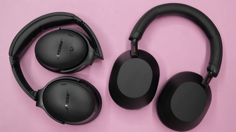 A pair of black Sony 1000XM5s and a pair of black Bose QC45s both laying face down on a pink surface.