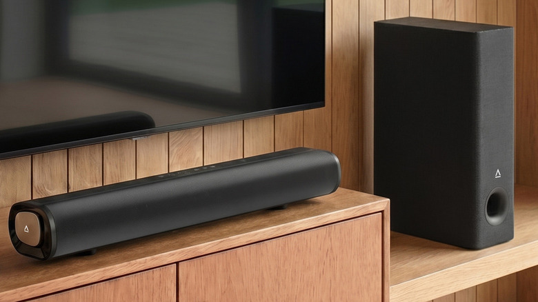 A soundbar placed on a wooden surface under a TV, next to a subwoofer.