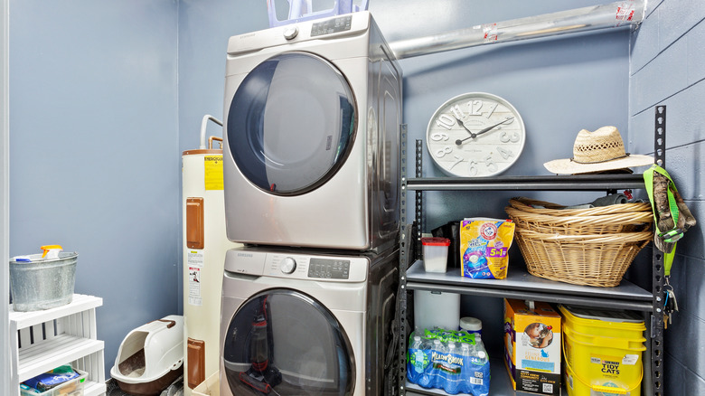A washer and dryer stacked in a small, blue room with a water heater and a shelf of household supplies