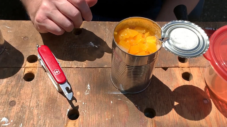 Swiss Army Knife with can opener tool exposed next to open can of fruit on wooden table