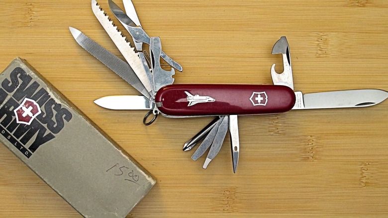 A Shuttle design Master Craftsman Victorinox Swiss Army Knife on a wooden table. All the tools are unfolded displaying blades, scissor, can opener and more. Next to the knife is the original box, which displays the words Swiss Army in large letters.