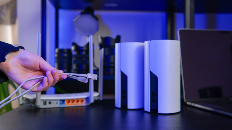 A person holding two ethernet cables next to wi-fi mesh nodes placed on a table alongside a router and laptop