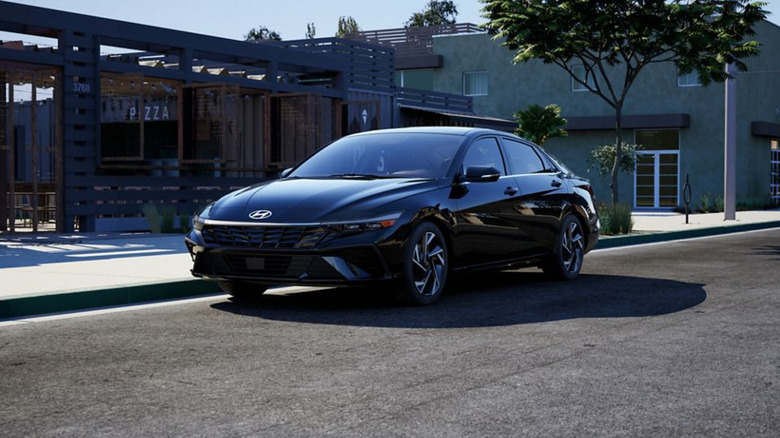 A black 2026 Hyundai Elantra sedan parked in the shade next to the pavement in front of a modern looking building.