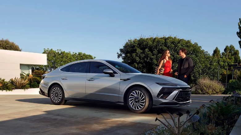 A silver 2026 Hyundai Sonata sedan standing in the driveway of a modern white house, with a couple standing on the other side of it.